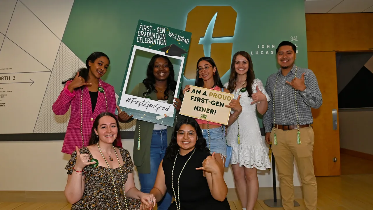 Group of students outside the Lucas Room in Cone Center.  They are smiling, holding picks up, with cords around their necks from First-Generation Graduation Celebration. One girl is holding up a foam frame sign that says "First-Generation Graduation Celebration - with a hashtag first gen grad." Another student is holding a sign that says, "I am a proud First-Generation Niner." 