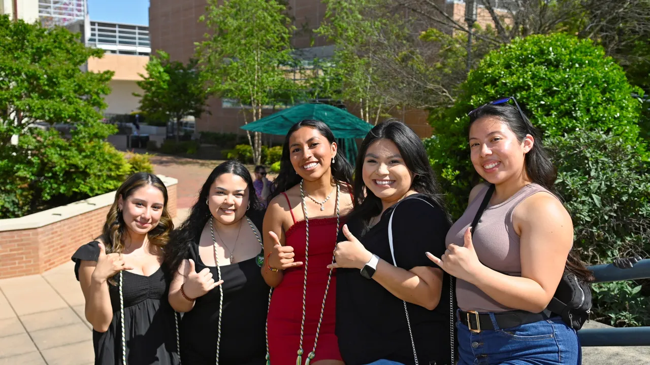 Group of women standing outside on stairs with cords hung around their necks, holding picks up, and smiling. There are 5 women, and the photo is from First-Generation Graduation Celebration.