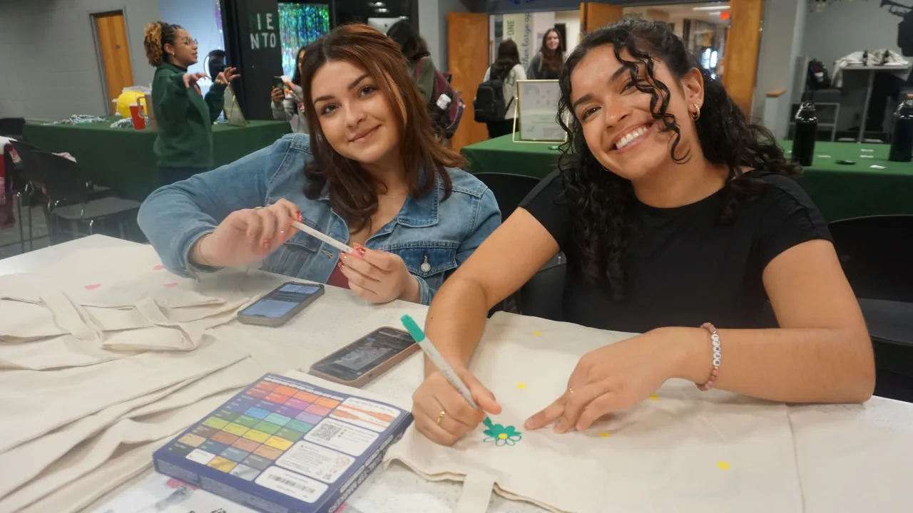 Two students sitting at a table and smiling during our Karaoke and Craft event in Spring 2025. They are both working on decorating tote bags.