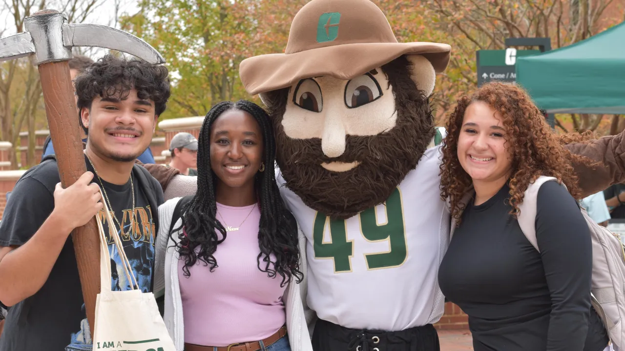 Three students standing and smiling with Norm the Niner mascot at a First-Generation event. One student is holding Norm's pickaxe and a canvas tote bag with the "I am a First Gen Niner" logo in Charlotte green. 