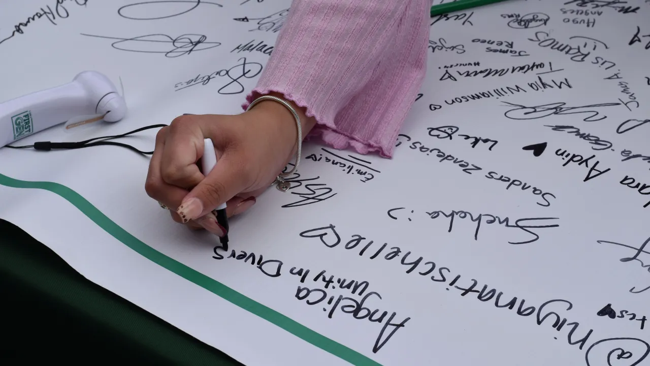Arm and hand of a student wearing a long sleeve, pink shirt and a bracelet signing the First-Gen Niner banner during First Gen Celebration Week.