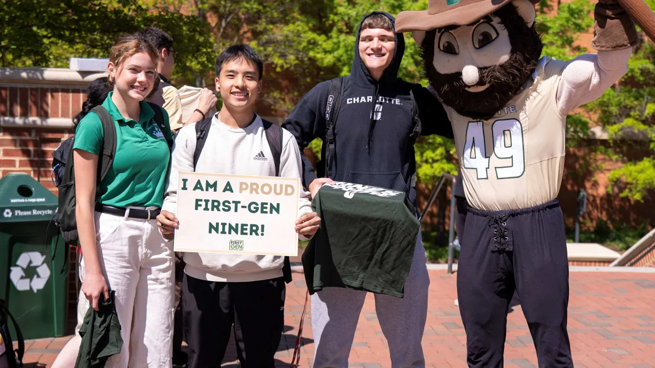 Three students standing and smiling with Norm the Niner mascot at a First-Generation Cookout. One student is holding a sign that says, "I am a pround First Gen Niner." Norm is holding up his pickaxe. They are all outside in Star Quad on a sunny day.