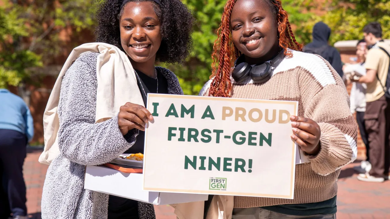 Two students smiling outside in Star Quad during First-Gen Appreciation Day. They are holding a sign together that says, "I am a proud First Gen Niner." 
