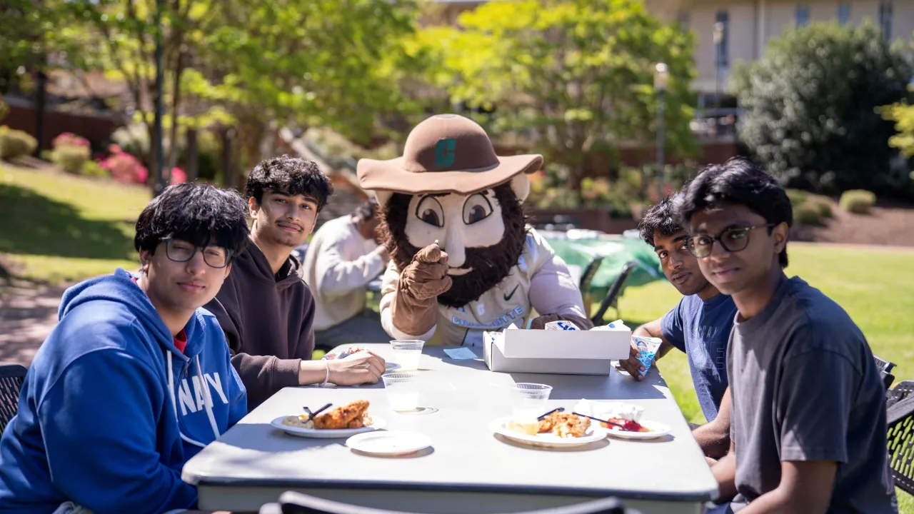 Group of four students and Norm the Niner at a table furing First-Gen Appreciation Day. The students have plates of food in front of them at the rectangular table. They are all looking at the camera, and Norm is pointing toward the camera as well. They are outside on a sunny day.