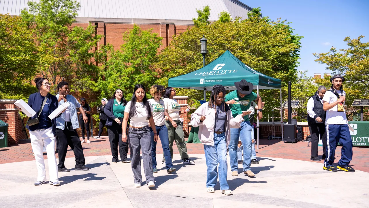 Group of people dancing in Star Quad at an event. There is a tent in the back with the logo for the office hosting the event. They are outside on a sunny day laughing and smiling. 