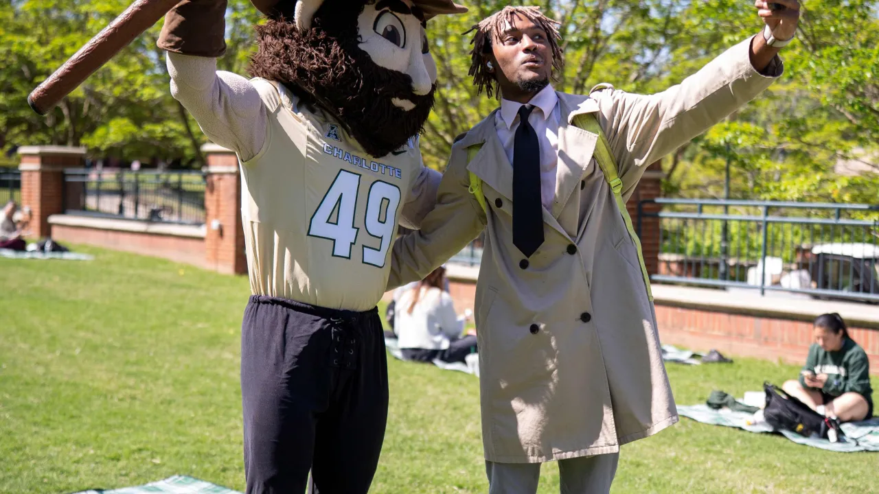 Student in a button up shirt, tie, and trench coat taking a selfie with Norm the Niner in Star Quad. There is a picnic blanket on the grass behind them, and Norm is raising his pickaxe into the air.