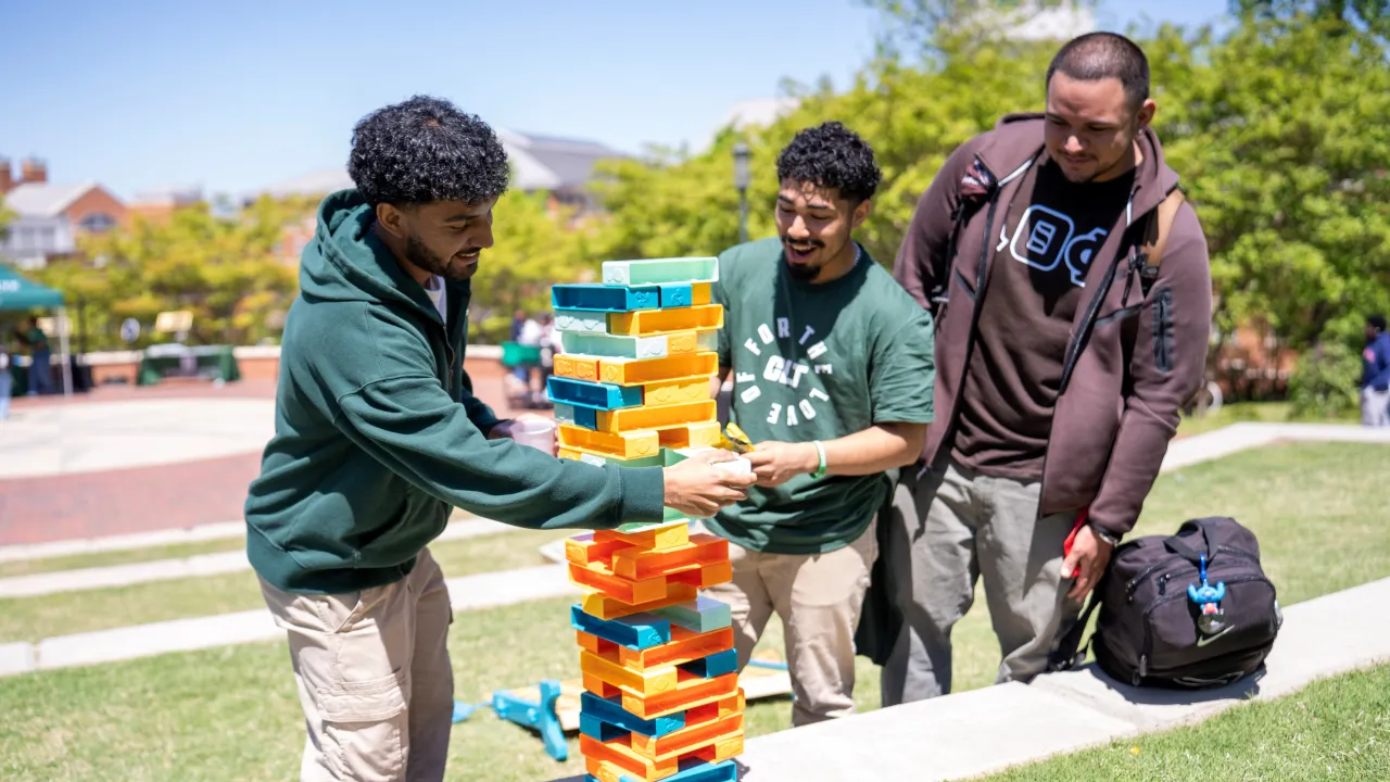 Three students outside during First-Gen Appreciation Day playing giant Jenga in Star Quad. One student is moving a piece from the tower, while the other two are leaned in watching.