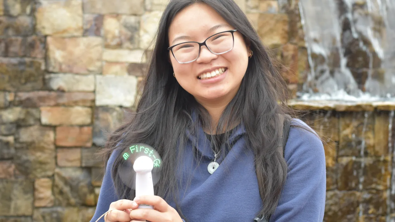 Student with glasses smiling in front of the fountain in Belk Plaza. She is holding a fan that is turned on and lights up with "First Gen" in green. 