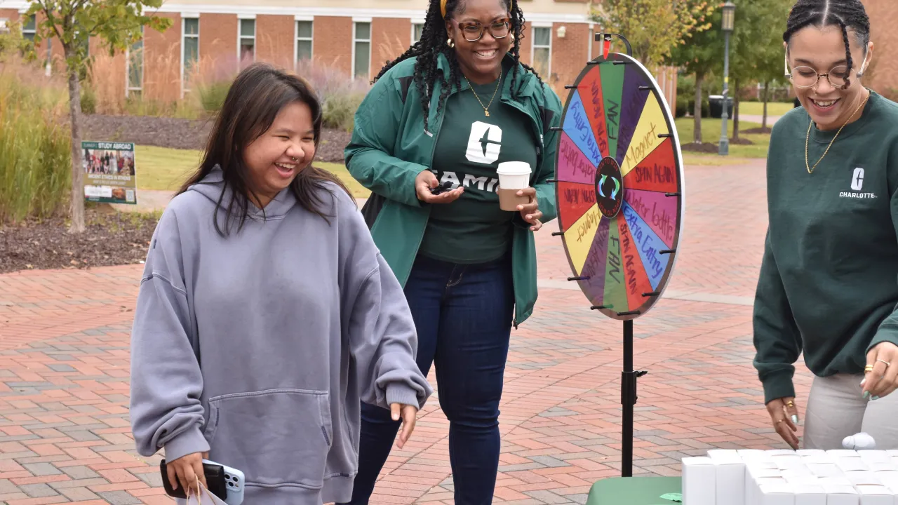One student laughing and smiling after spinning the prize wheel during Kickoff for First Gen Week. Two staff members are also around smiling and laughing while grabbing the prize for the student. They are all outside in the Belk Plaza.