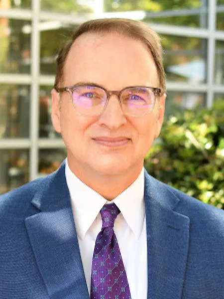 Man smiling softly facing forward wearing square glasses and a suit and tie. 