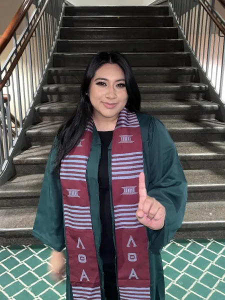 Woman holding her hand up in a number one sign standing by a staircase wearing a green graduation gown and greek stole