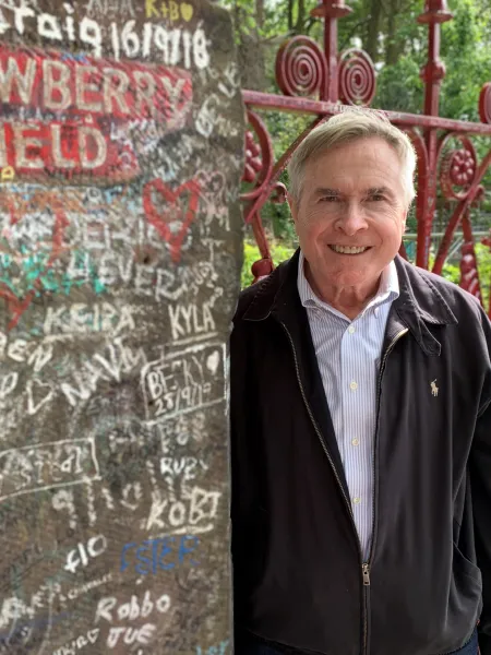 Man with light hair leaning against concrete wall wearing a button up shirt and a black coat