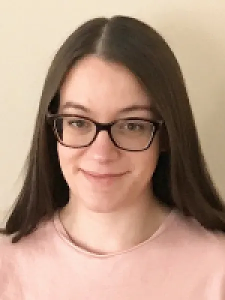 Woman with straight brown hair and glasses smiling in front of a beige background. She is wearing a pink, scoop neck top. 