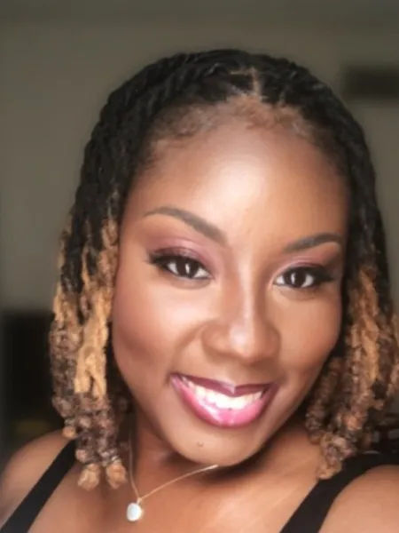 Woman smiling with her teeth looking sideways towards to camera, she has curly black and blonde hair 