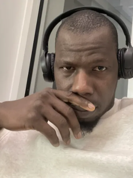 Black man with a buzzcut, black over-ear headphones and a heather gray crewneck. He is sitting in a building in front of a window and has his hand covering his mouth