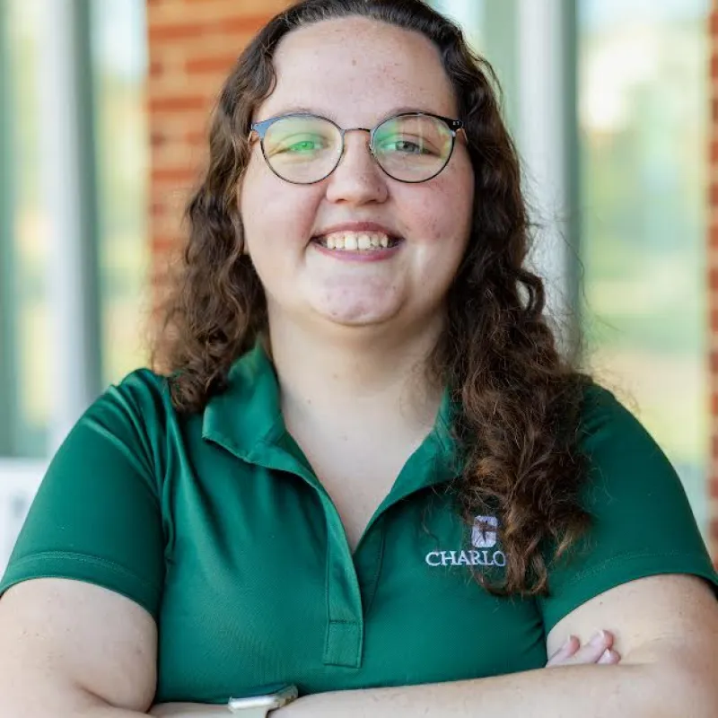 Woman with long, girly, brown hair wearing a green Charlotte polo with glasses and arms crossed. In the background, there is a brick building with windows