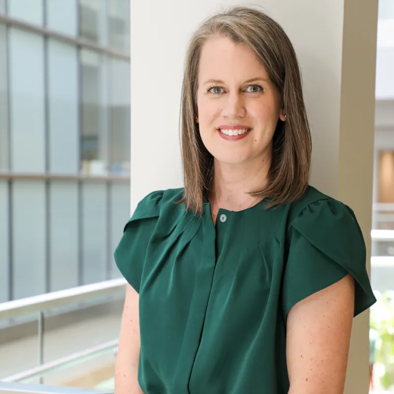 Woman with brown, shoulder-length hair smiling with a brick red lipstick. She is wearing a short sleeve deep green blouse and is standing in front of a pillar with windows in the background
