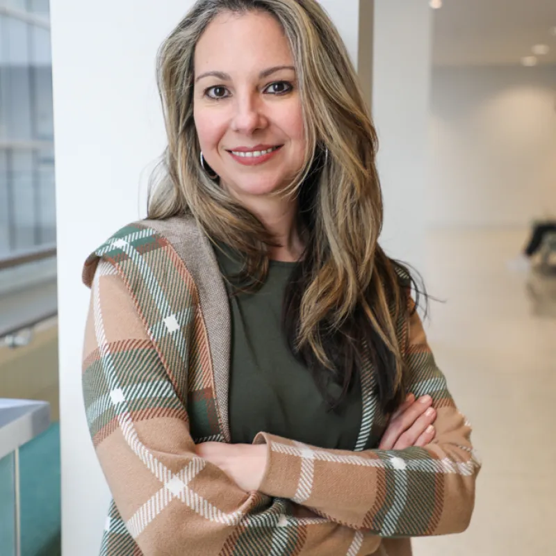 Woman with long brown and blonde hair standing in a building. Her arms are crossed, and she is smiling with teeth. She is wearing a green shirt with a earth-toned cardigan and black bottoms. 