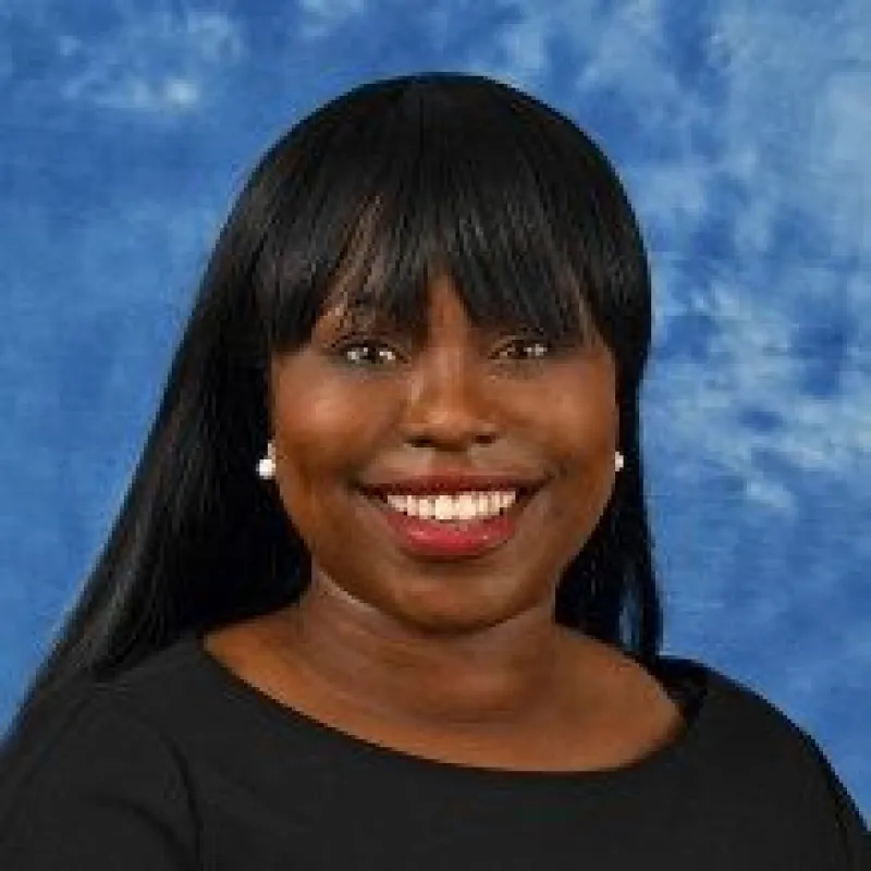 Black woman smiling in front of a blue background. She has long straight black hair with bangs, pearl earrings, and a black top on. 