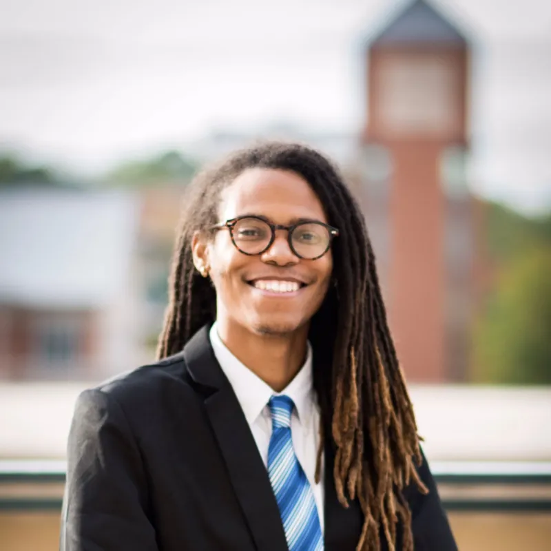 Black man with long locks and glasses smiling outside with buildings in the background. He is wearing a white dress shirt with a blue tie and a black blazer.