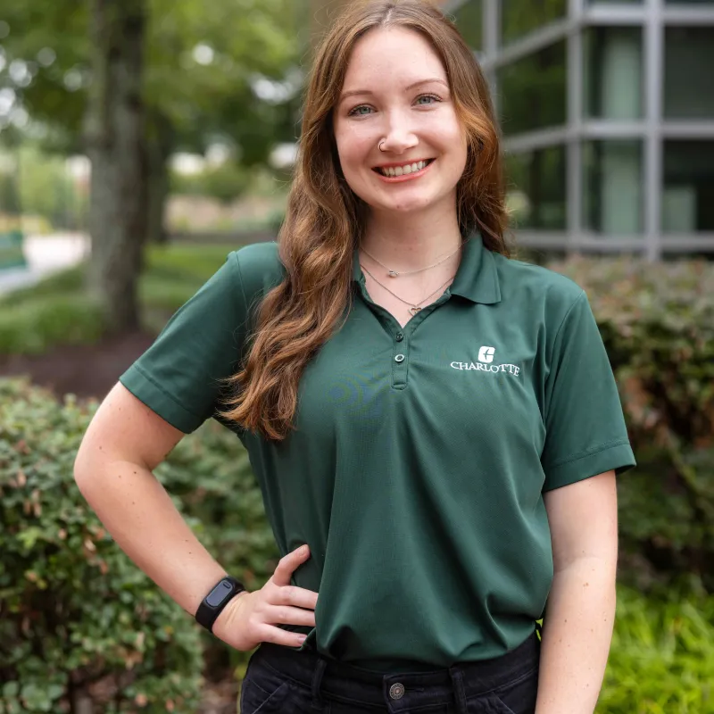 Person with long light brown hair standing outside with one hand on their hip. Smiling with a green polo and black jeans