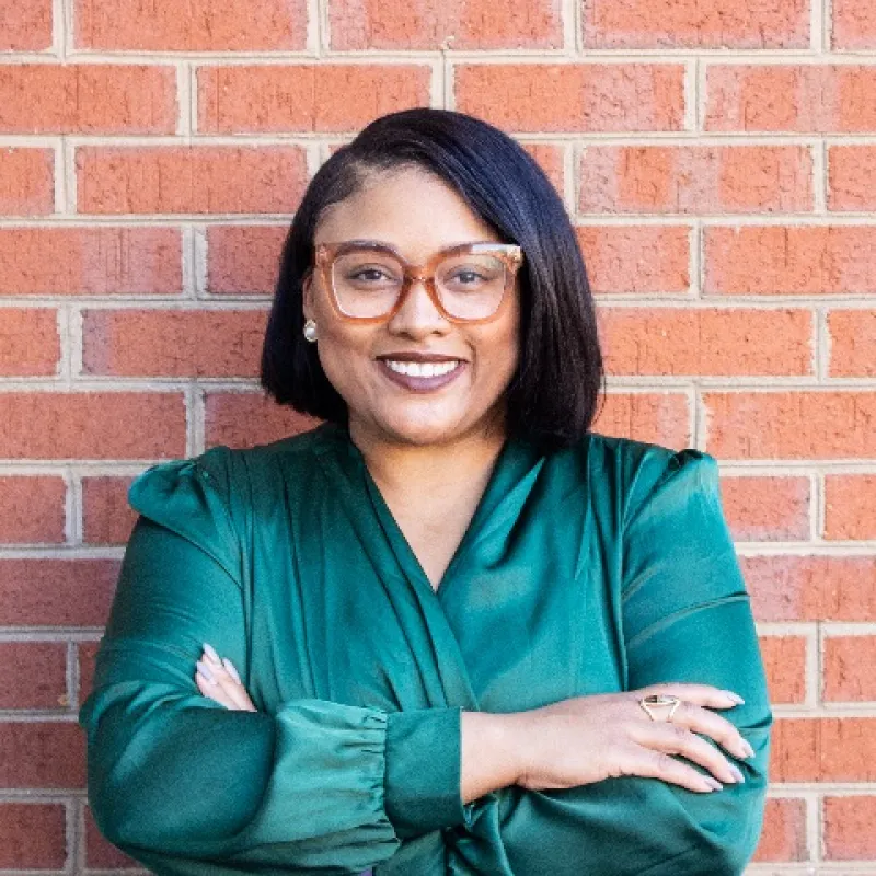 Woman in front of a brick wall wearing a green long sleeved blouse with arms crossed. Dark colored bob haircut, smiling, and wearing large framed brown glasses