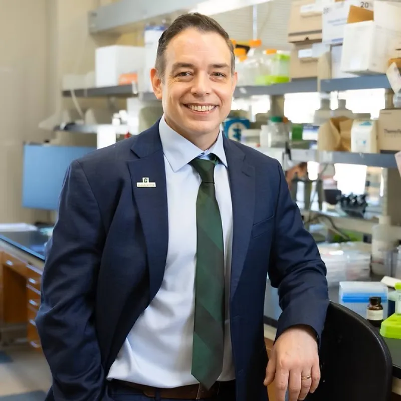 Man with a suit on smiling in a Biology lab. His suit is navy blue with a light blue button-up shirt with a Charlotte green tie. 
