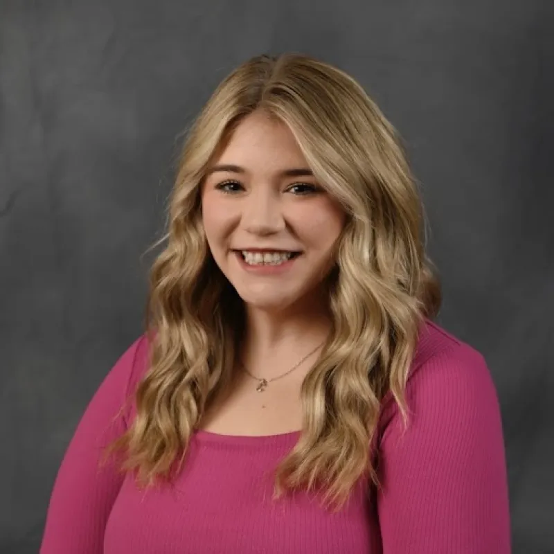 Woman with long blonde hair smiling with a pink long sleeve shirt and a necklace in front of a gray background