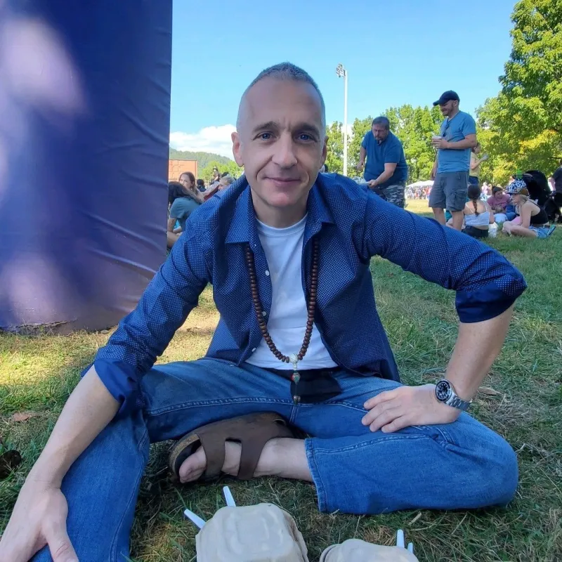 Man sitting on the ground wearing sandals, jeans, a white shirt, and an open blue button down shirt. One hand is on his leg and he is leaning forward and have other people in the background in an open green space