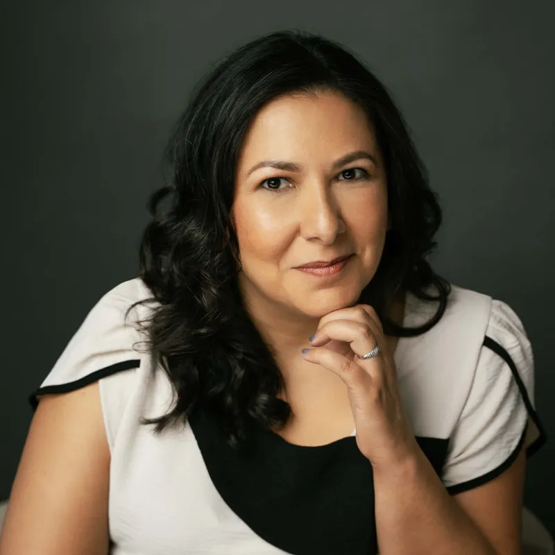 Woman with black hair in front of a dark gray background. She has on a black and white blouse, is sitting with her hand on her chin, and smiling without teeth.