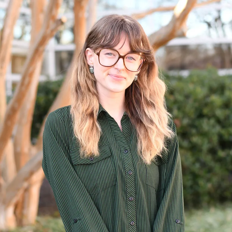 Person standing outside with a forest green button up shirt, large framed glasses, and curtain bangs. Their hair is ombre sandy brown to blonde and is medium length. they are also smiling without teeth.