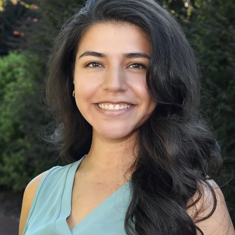 Smiling woman in a light blue sleeveless top and long, flowing, dark hair standing in front of trees