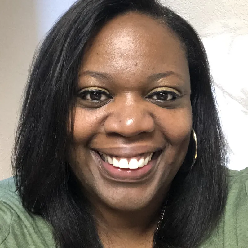 Black woman with medium length, black, straight, hair smiling, wearing hoop earrings, a gold necklace, and a green top.