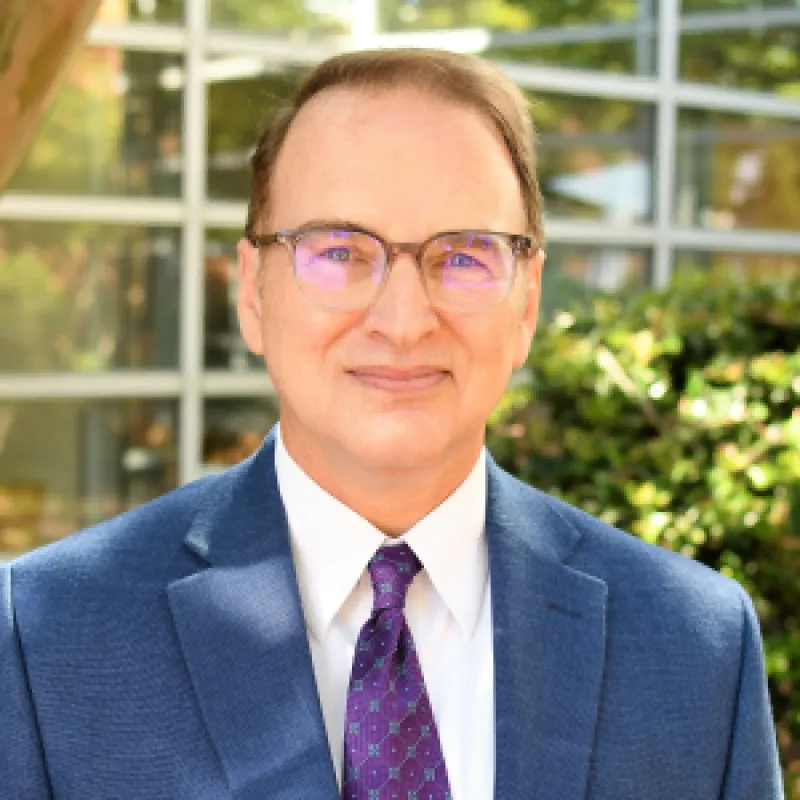 Man smiling softly facing forward wearing square glasses and a suit and tie. 