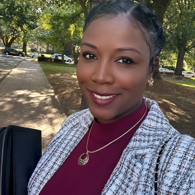 Brown-skinned woman smiling outside with trees and sidewalk in the background. She is wearing a maroon mock neck top with a black and white plaid blazer. Her necklace and earrings are gold, and she has her black hair tied back.