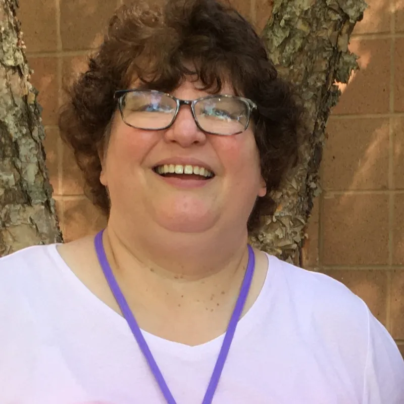 Woman with short curly hair and dark rimmed glasses smiling open mouthed. She's wearing a light pink shirt with a purple lanyard around her neck. In the background, there is a tree and a brick wall