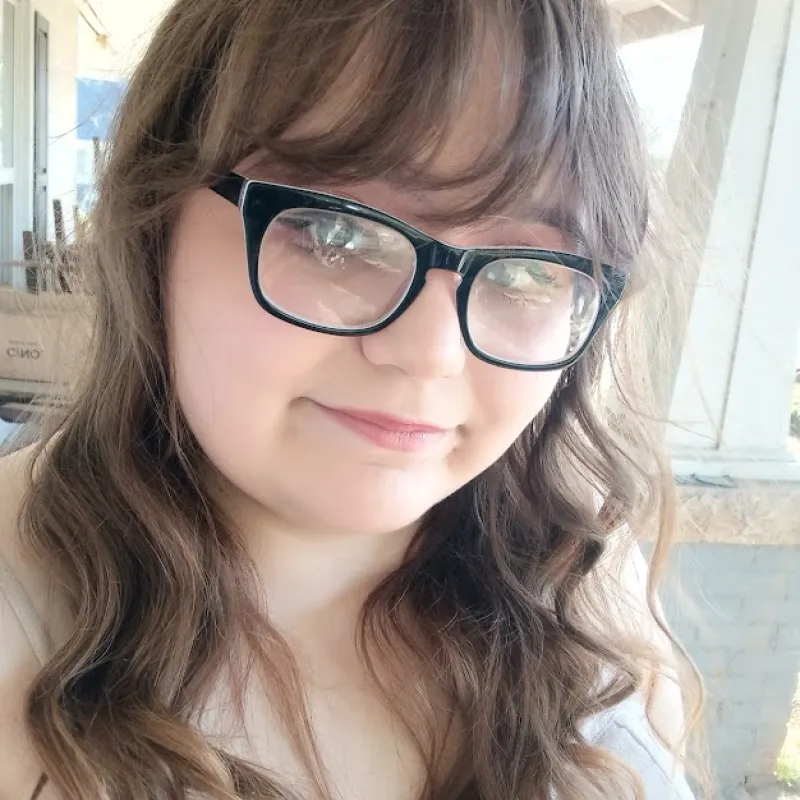 Women wearing black square glasses with wavy brown hair smiling softly at the camera