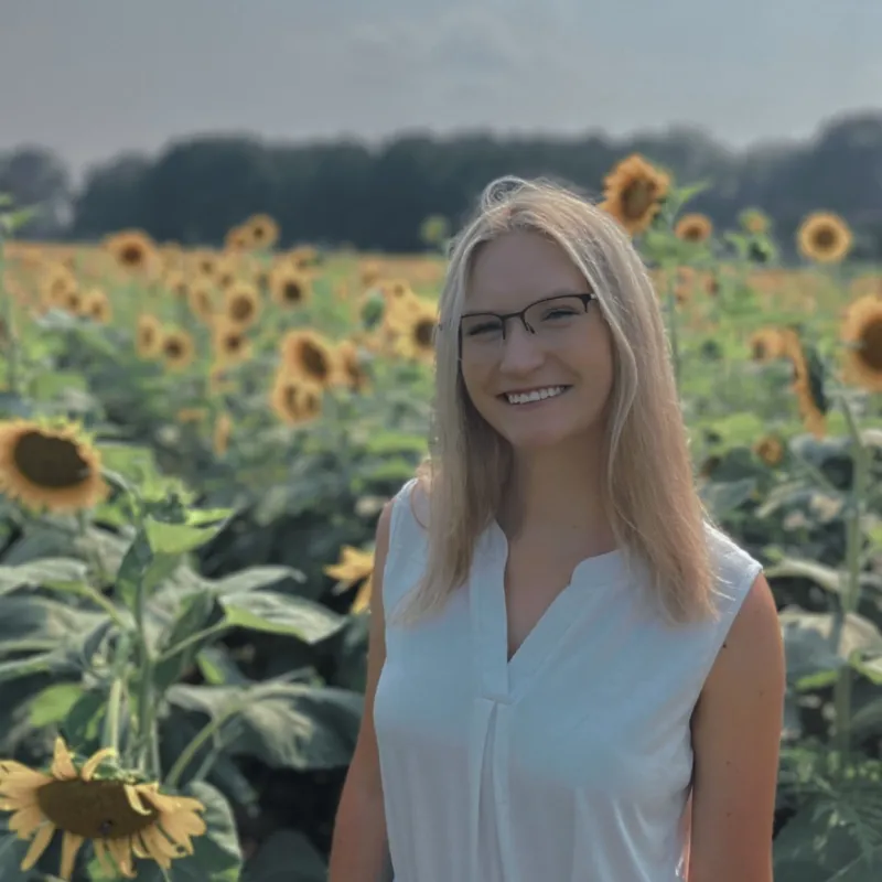 Blonde woman with black glasses and a white sleeveless shirt smiling in a field of sunflowers