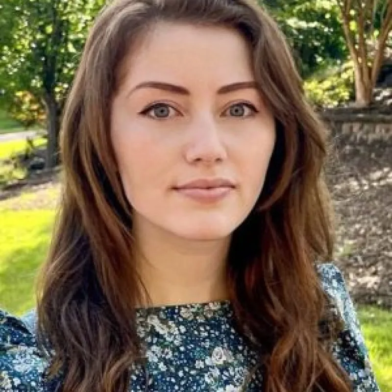 Woman with long brown hair wearing a floral blue blouse outside with tress and grass in the background