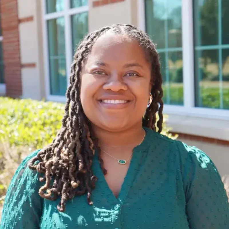 Black woman smiling in front of a brick building with medium-length wavy locs, hoop earrings, and a necklace. She is wearing a deep green, textured blouse.