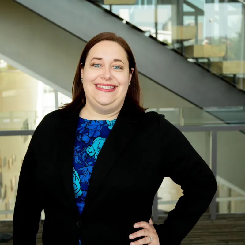Woman with a blue top and a black cardigan smiling in front of some glass windows and a stairwell. She has red lipstick, straight brown hair, and her hand on her hip