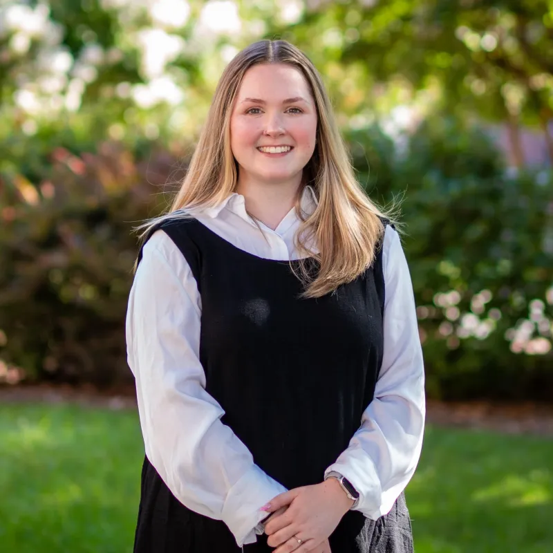 White woman with blonde hair smiling outside with a whit button down shirt under a black sleeveless dress
