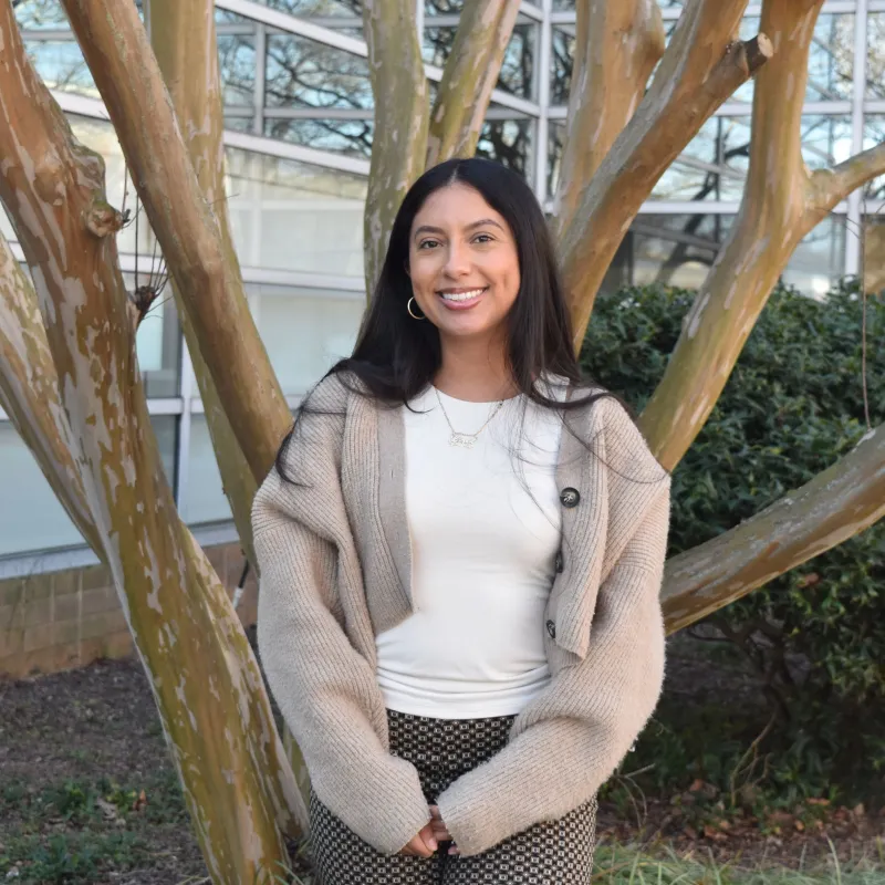 Person with long dark hair smiling outside in front of a tree. They have a light brown cardigan and a white shirt