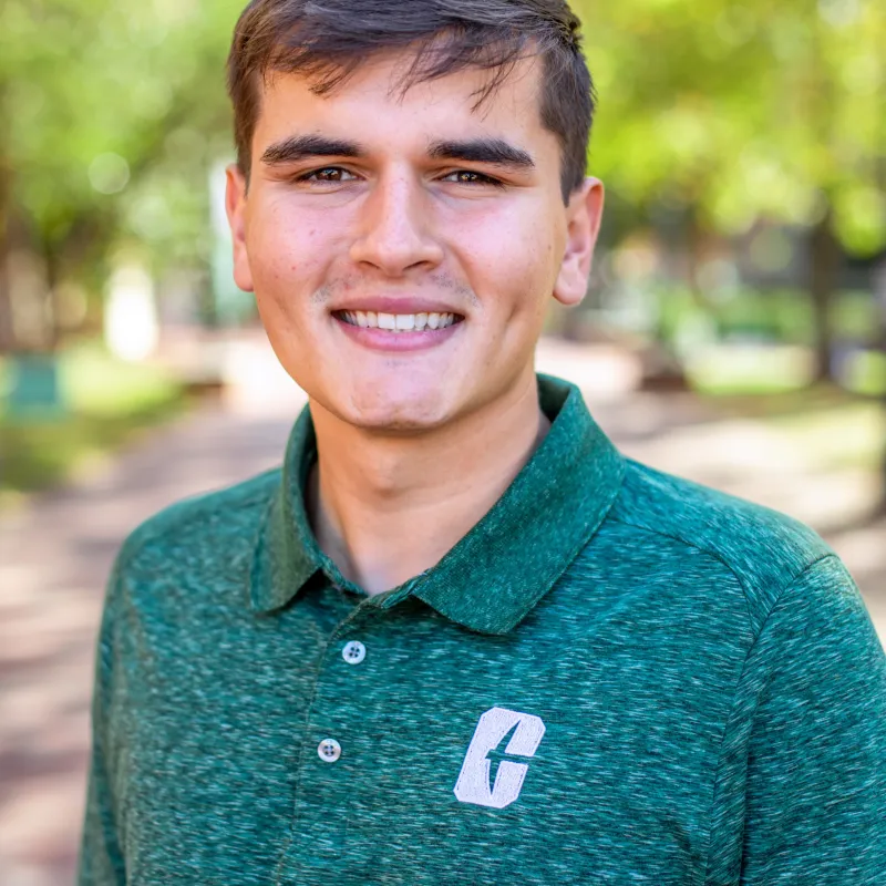 Young man with brown short hair and dimples wearing a green Charlotte polo shirt smiling outdoors with a walkway and trees in the background