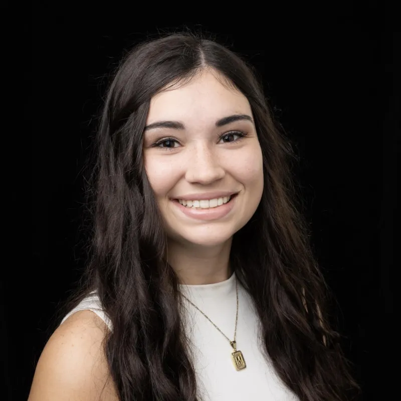 Woman with light skin and dark long hair. she is smiling in front of a black background and wearing a white sleeveless top with a gold necklace