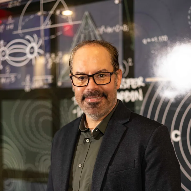 man with glasses, short brown hair and a full beard smiling. he is wearing a green button down shirt and a blazer with his hands in pockets