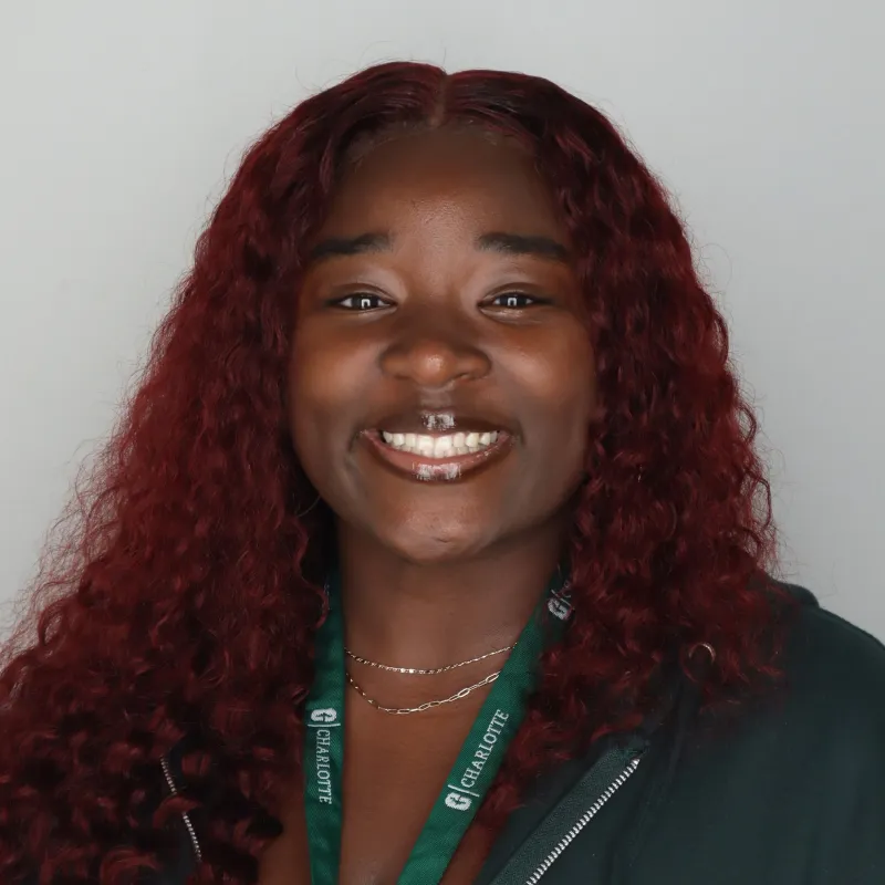 Woman with black cherry colored hair smiling in front of a plain white background. She has a Charlotte branded zip-up hoodie and lanyard on. Both are green.