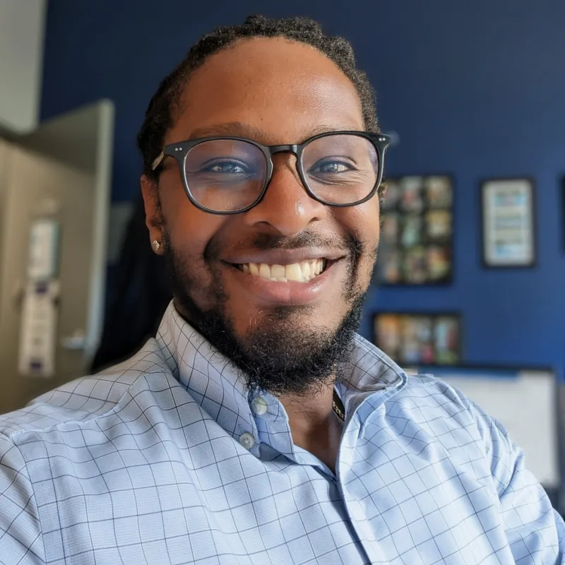 Man smiling in an office with blue walls. He has black glasses, a beard, and a blue button-down shirt on.