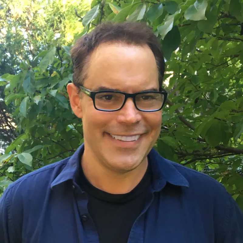 Man with square glasses and navy blue button up smiling at camera with green trees in background