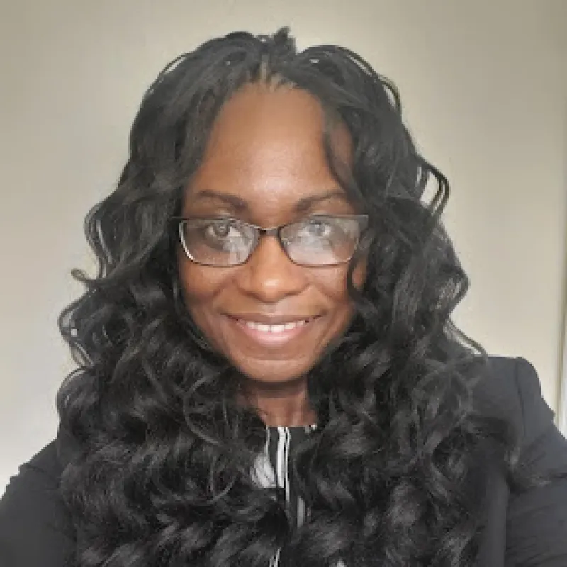 Black woman with long, black, wavy hair and black rectangular glasses smiling and wearing black professional blazer. 